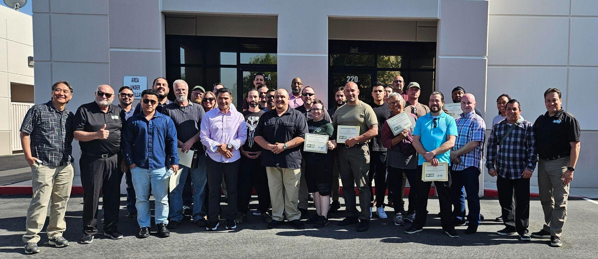 A diverse group, including Las Vegas security team members, stands outside a building holding certificates and smiling after completing the antonio-leadership-profile program, celebrating their achievement in casual attire.