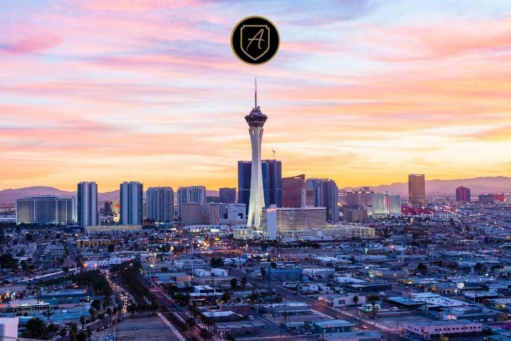 The Las Vegas skyline at sunset, with the Stratosphere Tower and city buildings beneath vibrant pink, orange, and purple clouds. Centered above is a black circle with a gold.