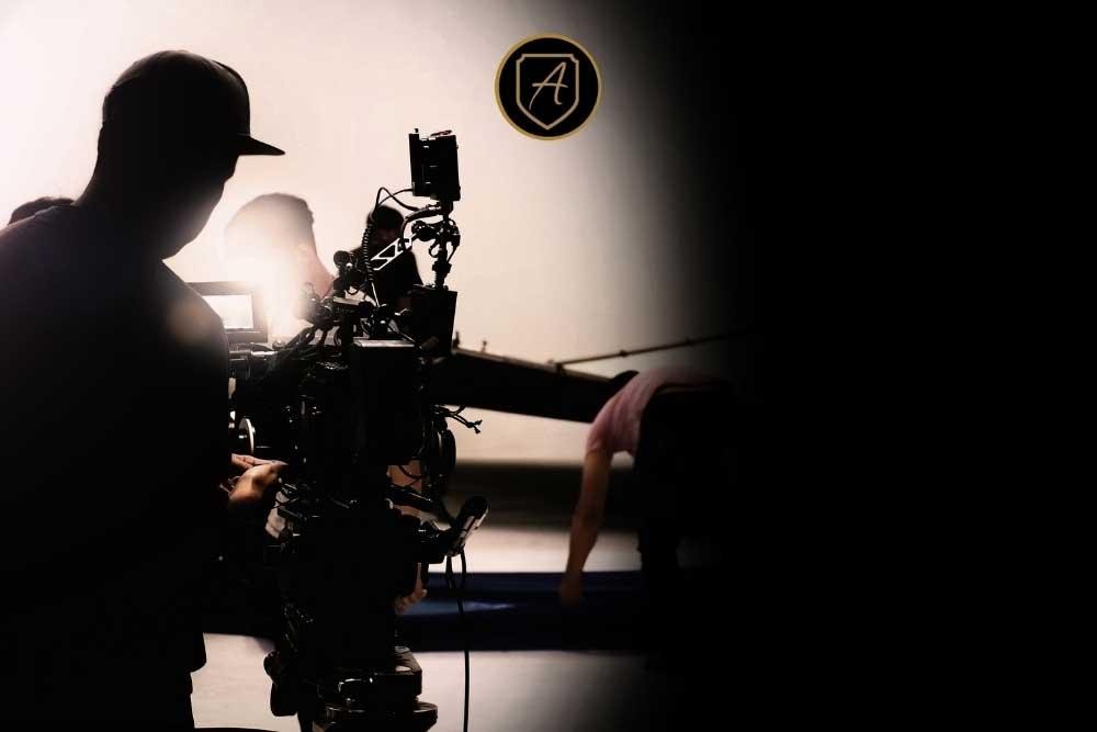 A silhouetted camera operator films under bright set lights while another crew member adjusts equipment. The film-set-security-las-vegas1 logo, featuring a circular A, appears at the top center of the scene.