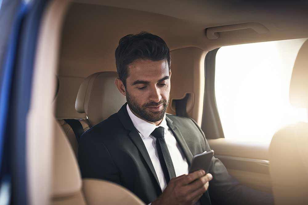A man in a suit relaxes in the beige interior of secure-ground-transporation-4, enjoying Nevada sunlight through the window as he checks his smartphone, experiencing premium comfort and safe ground travel.