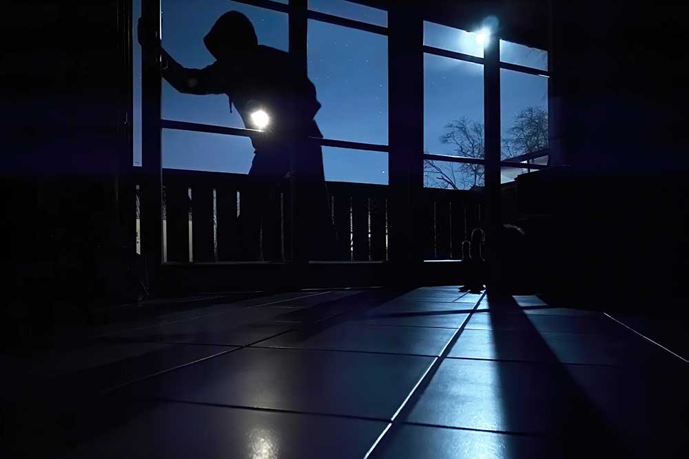 A hooded figure attempting a nighttime break-in through a glass door, symbolizing modern security threats handled by Alexios Security Solutions.