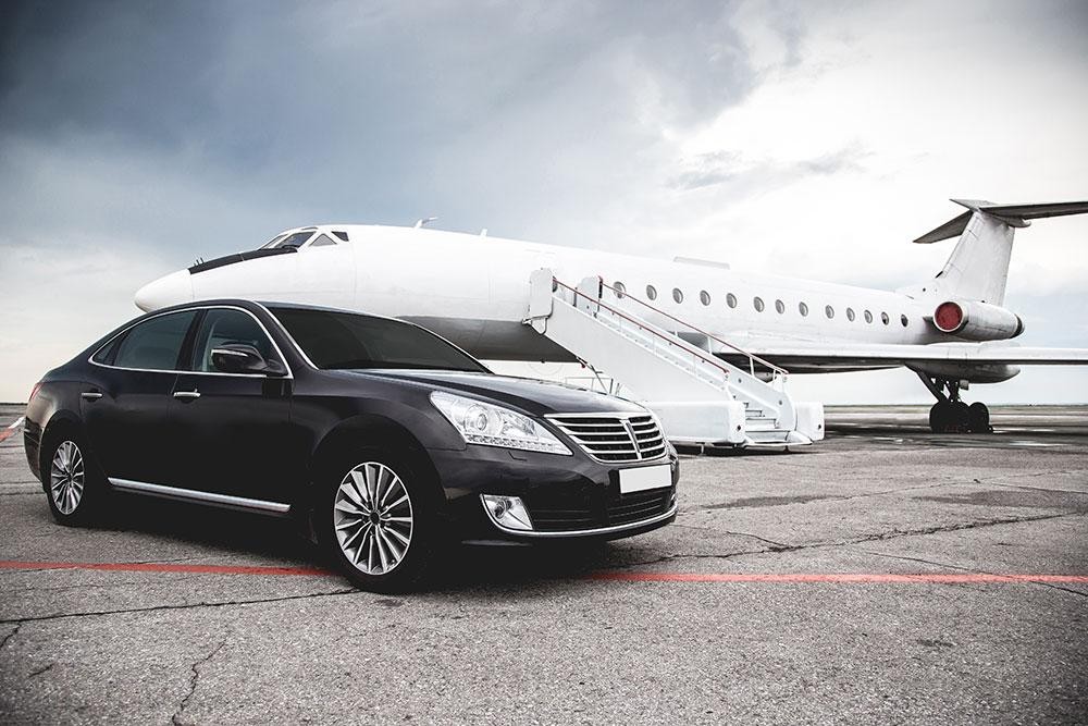 The Executives-Need-Secure-Ground-and-Air-Transportation-2 features a black luxury sedan on an airport tarmac beside a white private jet with staircase access under a cloudy sky.