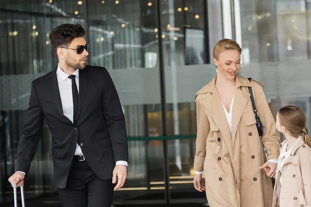 A man in a black suit and sunglasses, representing executive-protection-page-5 by Alexios Security Solutions in Las Vegas, pulls a suitcase beside a woman in a beige trench coat and a smiling girl outside a modern building.