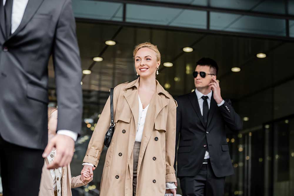 A smiling woman in a beige trench coat exits a modern glass building with a child and two men in suits—one adjusting his sunglasses—demonstrating Executive Protection Services in Las Vegas and Phoenix by Alexios Security. A smiling woman in a beige trench coat exits a modern glass building with a child and two men in suits—one adjusting his sunglasses—demonstrating Executive Protection Services in Las Vegas and Phoenix by Alexios Security.