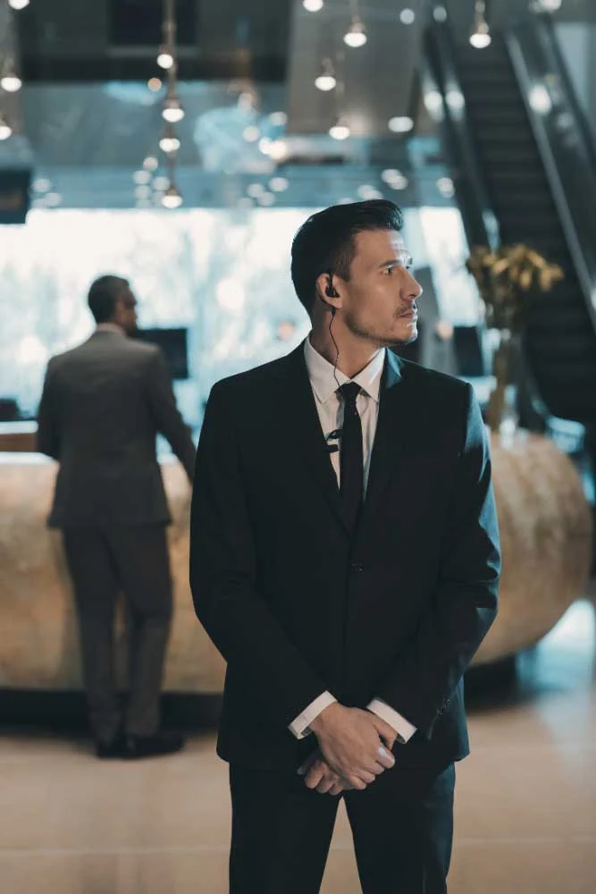 A man in a suit with an earpiece stands alert in a lobby, showcasing medium-carousel-4 by Alexios Security Solutions for Executive Protection & Bodyguards in Las Vegas, while another suited man is at a counter near an escalator.