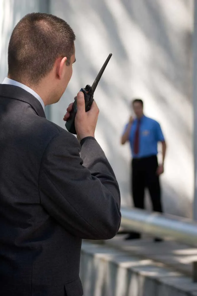 A man in a suit with a walkie-talkie stands in the foreground, another on the phone nearby—highlighting medium-carousel-5 by Alexios Security Solutions for Executive Protection & Bodyguards in Las Vegas outside a white building.
