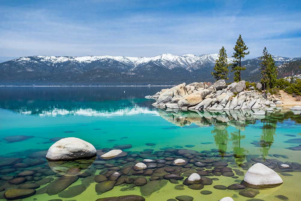 Alexios-security-solutions-lake-tahoe-nevada: Turquoise lake with smooth rocks, pine trees on rocky shores near Reno, and distant snow-capped mountains beneath a blue sky. Alexios-security-solutions-lake-tahoe-nevada: Turquoise lake with smooth rocks, pine trees on rocky shores near Reno, and distant snow-capped mountains beneath a blue sky.