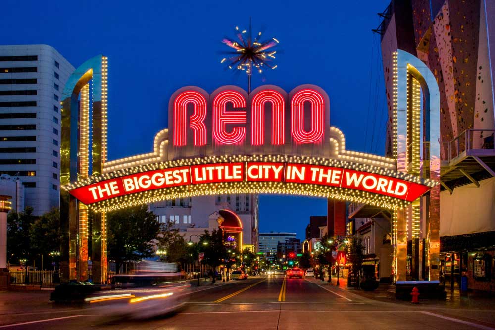 At night, the Reno arch glows with bold red RENO letters and a lit sign reading THE BIGGEST LITTLE CITY IN THE WORLD, as buildings and traffic bustle below—protected by Alexios-security-solutions-reno-nevada.