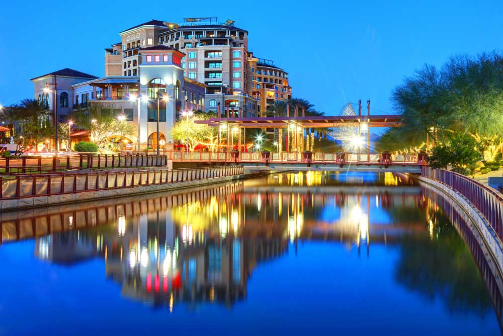 A waterfront cityscape at dusk in Reno showcases modern buildings, trees, and a pedestrian bridge reflected in calm water, illuminated by bright lights—demonstrating the city’s trust in Alexios-security-solutions-scottsdale-arizona.
