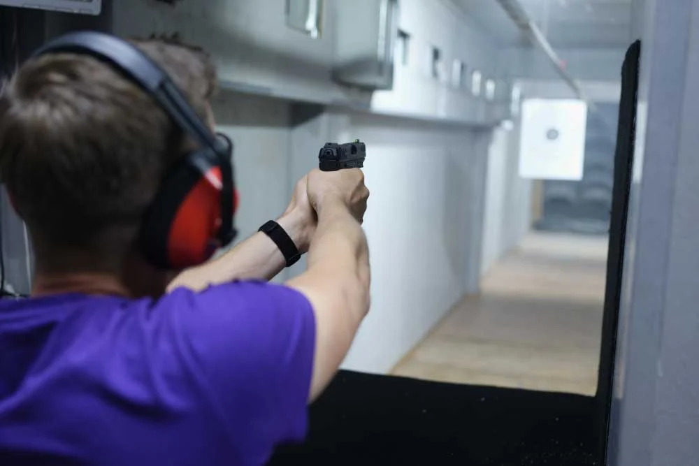 A person in a purple shirt wearing red and black ear protection aims a handgun at a paper target during a CCW-training-class-image firearm safety session at an indoor range in Las Vegas. | Executive Protection & Bodyguards in Las Vegas | Alexios