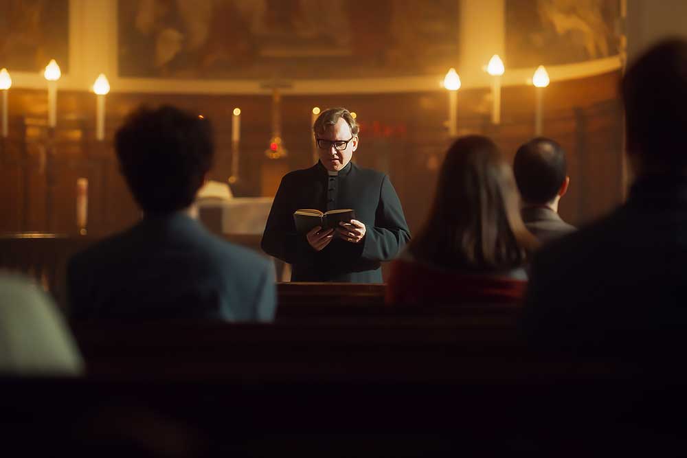 A priest in black robes reads from a book at the front of a church in Arizona, candles glowing behind him. The scene highlights safety with Security-for-Houses-of-Worship-in-Nevada-and-Arizona-1. A priest in black robes reads from a book at the front of a church in Arizona, candles glowing behind him. The scene highlights safety with Security-for-Houses-of-Worship-in-Nevada-and-Arizona-1.