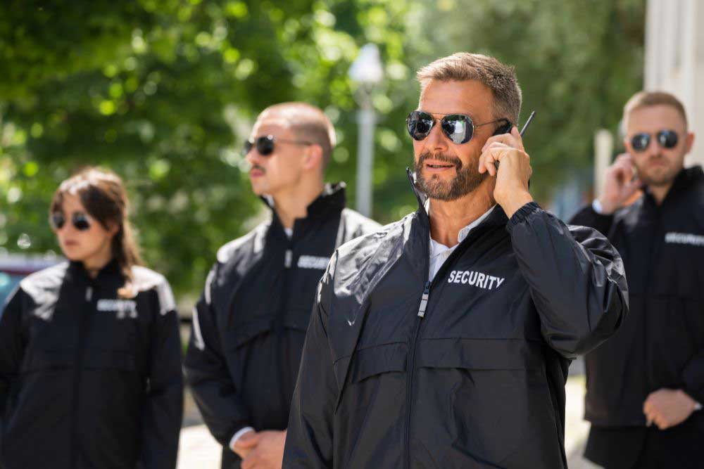 Four uniformed guards from Uniformed-Security-Guard-Services-1, dressed in black jackets and sunglasses, stand alert outside. One speaks into an earpiece as the others scan the area, showcasing professional security and vigilance. Four uniformed guards from Uniformed-Security-Guard-Services-1, dressed in black jackets and sunglasses, stand alert outside. One speaks into an earpiece as the others scan the area, showcasing professional security and vigilance.