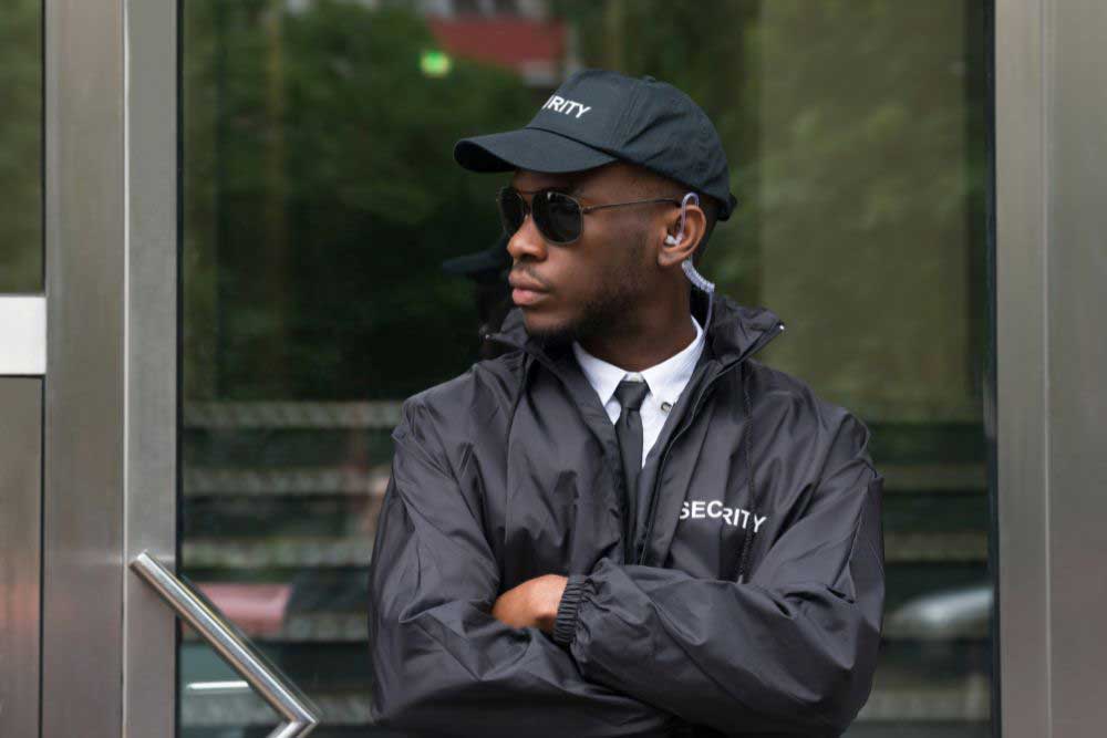 A uniformed security guard in a black jacket, cap, and sunglasses stands with arms crossed before a glass door, attentively watching the surroundings—showcasing Uniformed-Security-Guard-Services-2. A uniformed security guard in a black jacket, cap, and sunglasses stands with arms crossed before a glass door, attentively watching the surroundings—showcasing Uniformed-Security-Guard-Services-2.