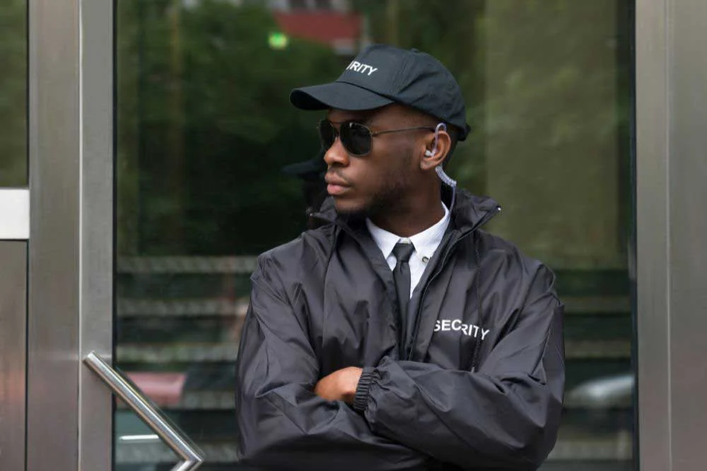 A uniformed security guard in a black jacket, cap, and sunglasses stands with arms crossed before a glass door, attentively watching the surroundings—showcasing Uniformed-Security-Guard-Services-2.