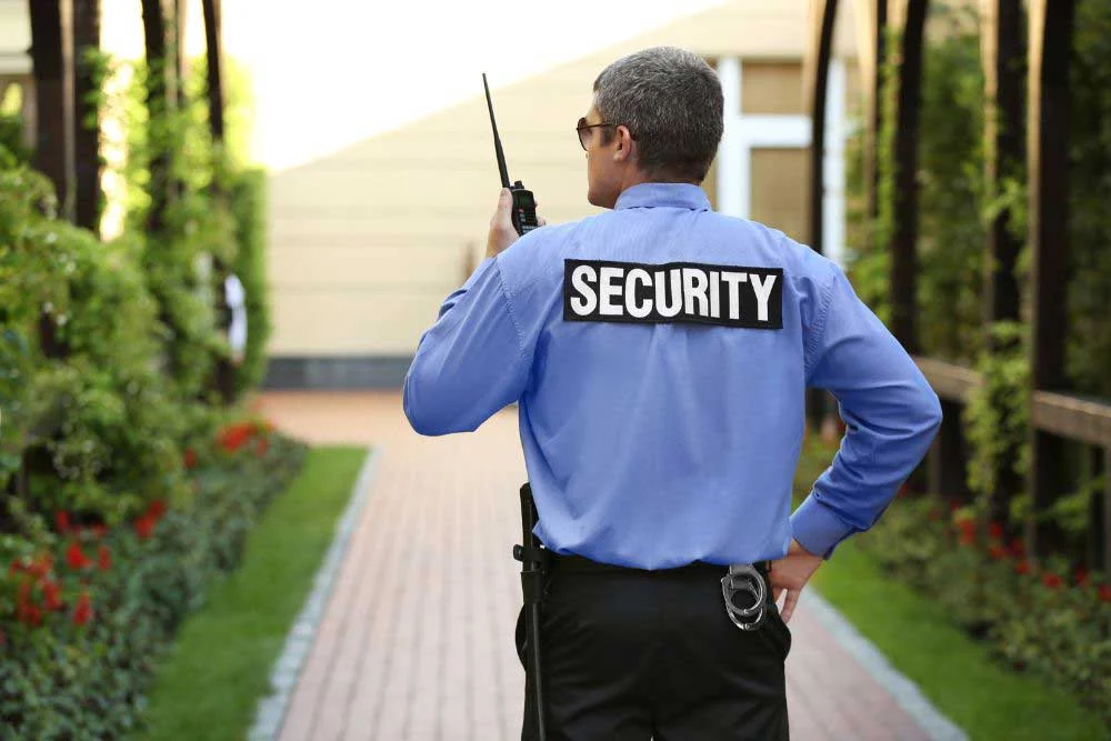 A Uniformed-Security-Guard-Services-6 officer in blue with SECURITY on the back stands on a path, walkie-talkie in hand, surrounded by greenery and flowers, facing away to ensure safety in this peaceful setting.