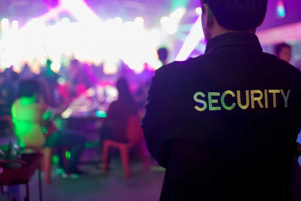 A Uniformed-Security-Guard-Services-7 officer in Nevada stands with their back to the camera, supervising a lively, well-lit event where colorful lights shine and guests are seated at tables in the background.