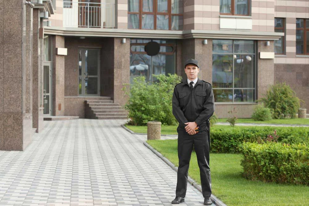 A residential security guard in a black uniform stands on a tiled walkway before a modern building, ensuring estate safety with estate-and-asset-protection-3.