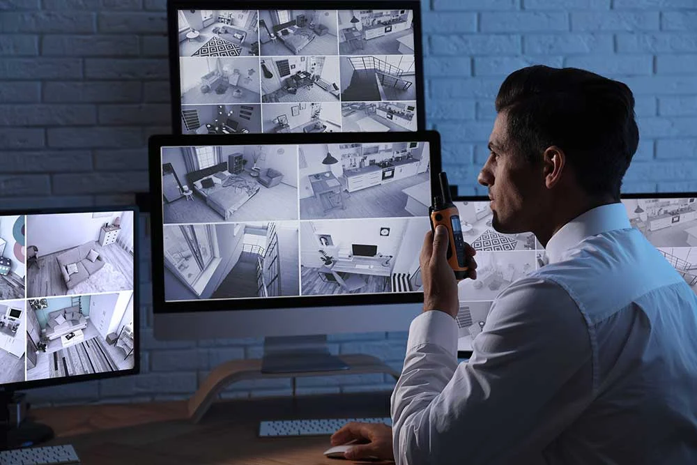 A security guard in a white shirt uses estate-and-asset-protection-5 and a walkie-talkie to monitor multiple surveillance cameras on computer screens, ensuring executive home security in a dimly lit room.