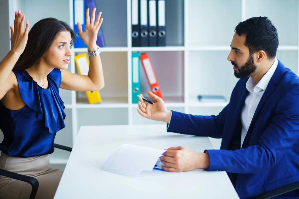 During a hostile-termination-training-1 session in Arizona, a frustrated woman raises her arms while discussing with a man in a business suit at an office desk surrounded by documents and binders.