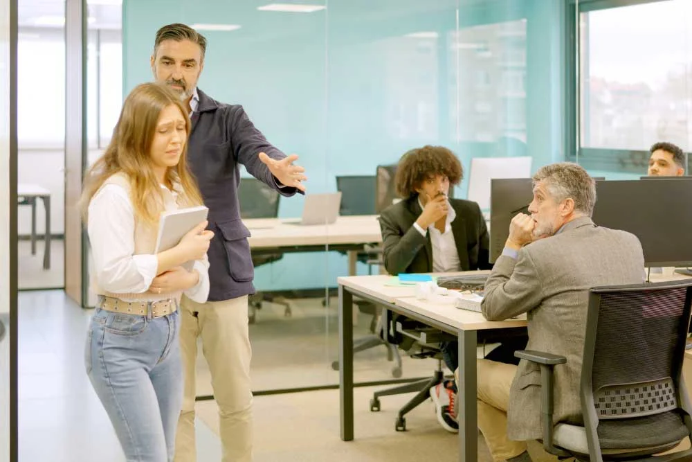 As a man gestures toward the door, an upset young woman with a laptop leaves, while three colleagues observe from their desks in a modern glass-walled office—highlighting the need for employee separation security like hostile-termination-training-2.
