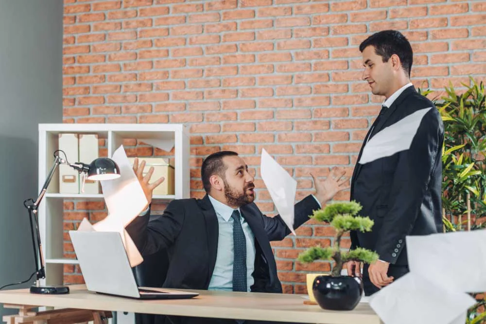 Two men in suits argue in an office, with one angrily throwing papers. This shows the importance of using hostile-termination-training-6 for workplace violence training and secure employee separation to keep offices safe.