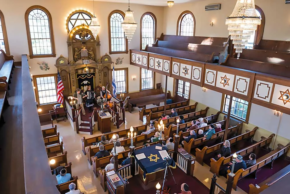 A top-down view of a synagogue interior during service, showing congregants, pews, stained glass, chandeliers, Jewish symbols like Stars of David, and house-of-worship-security-1 measures in place throughout.