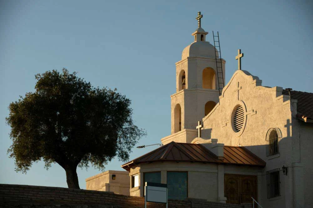 Basking in warm Nevada sunlight, a beige stucco church with three crosses and a bell tower stands by a large tree, embodying the serenity and security provided by house-of-worship-security-2.