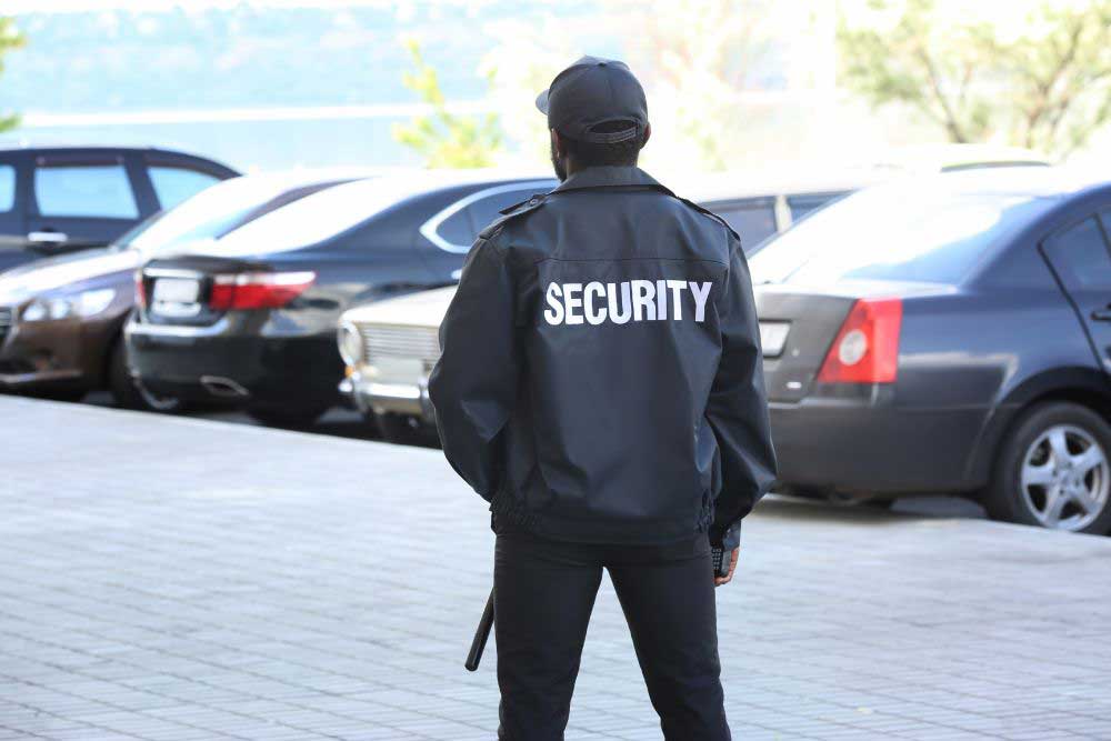 A security guard wearing the house-of-worship-security-4 black jacket labeled SECURITY stands in a parking lot with parked cars, protecting the church and ensuring safety while facing away from the camera. A security guard wearing the house-of-worship-security-4 black jacket labeled SECURITY stands in a parking lot with parked cars, protecting the church and ensuring safety while facing away from the camera.