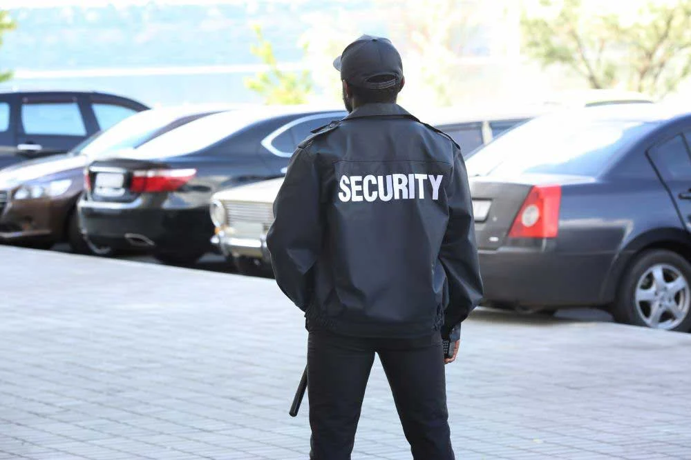 A security guard wearing the house-of-worship-security-4 black jacket labeled SECURITY stands in a parking lot with parked cars, protecting the church and ensuring safety while facing away from the camera.