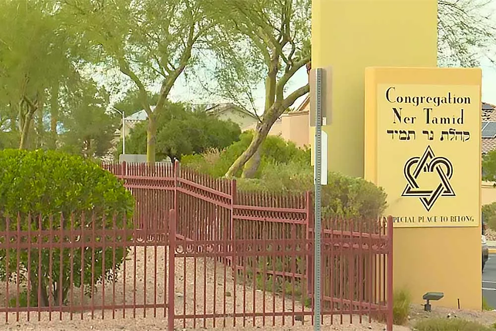A yellow house-of-worship-security-5 sign with Hebrew text and a Star of David stands by the entrance of Congregation Ner Tamid near a red fence, reflecting increased security for houses of worship.