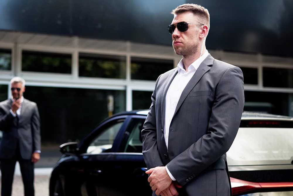 Two men in suits and sunglasses provide Secure-Ground-Transportation-2 near a black car outside a modern Nevada building. One man stands with hands clasped, while the other communicates via earpiece.