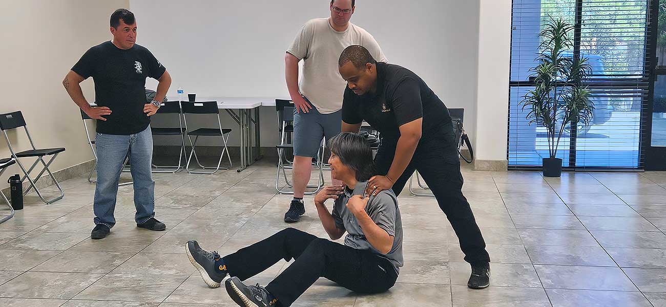 In a room with chairs, a table, and large windows, four people demonstrate Protection-Response-Tactics-1 on tile floors: one sits while another instructs using their arms; two others observe nearby. In a room with chairs, a table, and large windows, four people demonstrate Protection-Response-Tactics-1 on tile floors: one sits while another instructs using their arms; two others observe nearby.