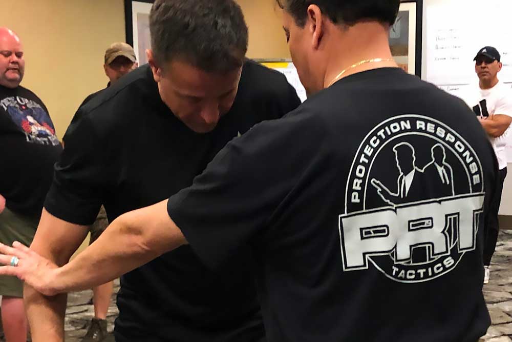 Two men practice self-defense indoors as one guides the other's arm down. The man facing away wears a "Protection-Response-Tactics-4" shirt, demonstrating effective techniques while several onlookers watch in the background.