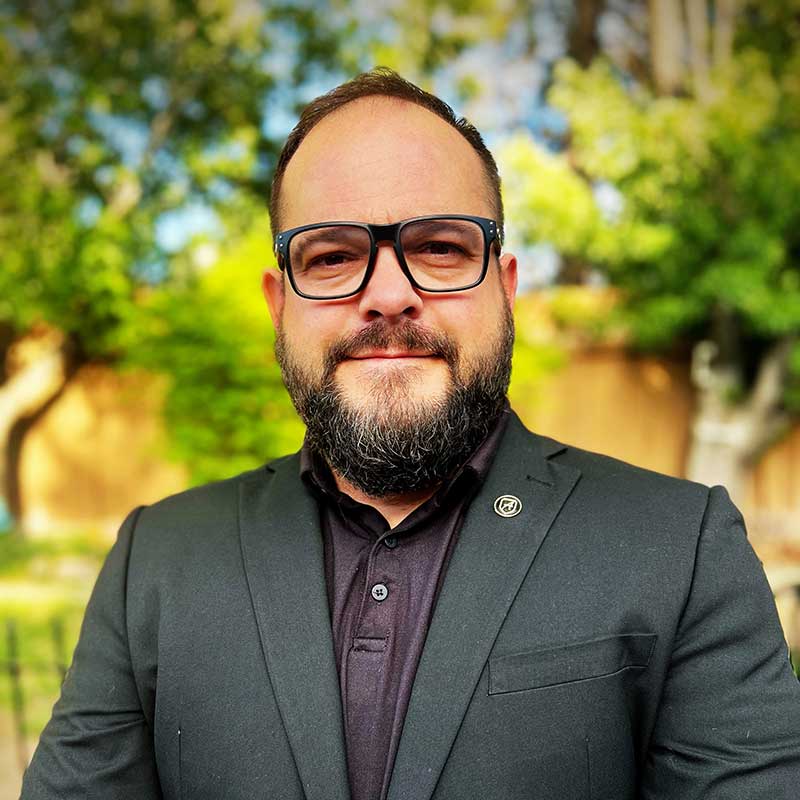 Richard Garretson, a bearded man in glasses, a black suit jacket, and a black polo shirt, stands outdoors before blurred trees and greenery, smiling at the camera—representing Nevada security services. | Executive Protection & Bodyguards in Las Vegas | Alexios