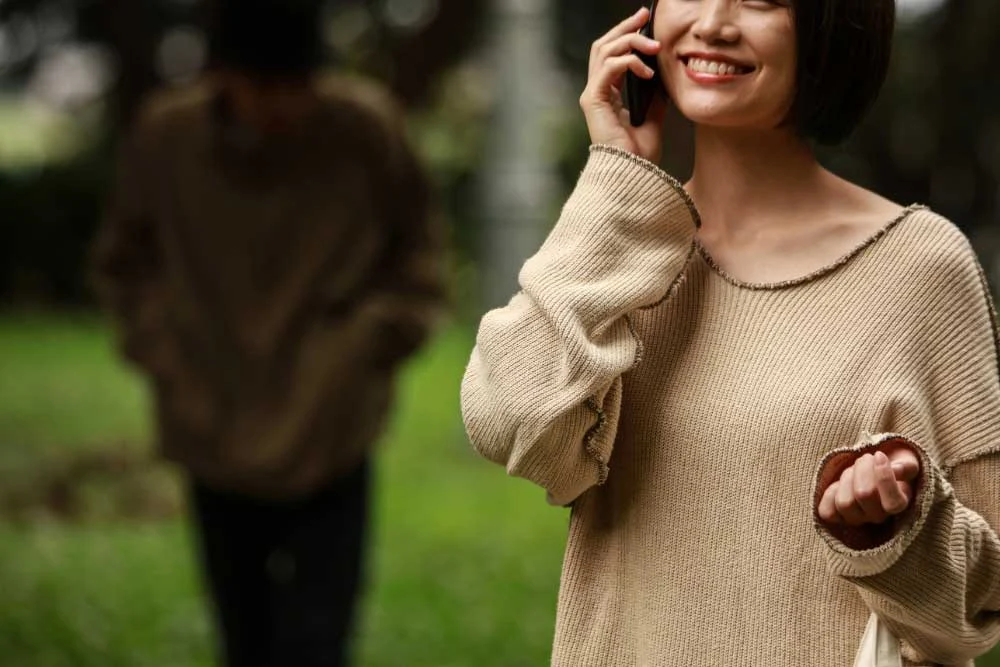 A woman in a beige sweater smiles while talking on her phone outdoors, staying alert with stalker-detection-and-prevention-1, as a blurred figure stands on the grass in the background.