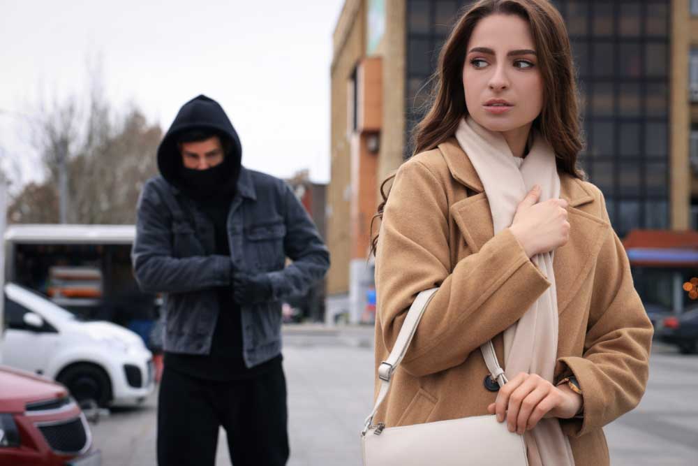 In an urban setting, a woman in a tan coat and white scarf looks over her shoulder with concern as stalker-detection-and-prevention-2 alerts her to a hooded man in dark clothing approaching from behind.