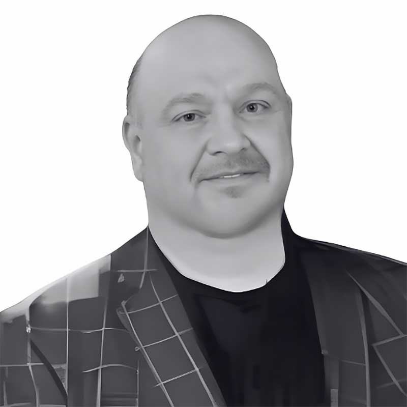 A man with a bald head and mustache, wearing a dark checkered jacket over a black shirt, smiles slightly against a plain white background—featured in the Antonio Guadagni-headshot product as an executive protection expert in Nevada.
