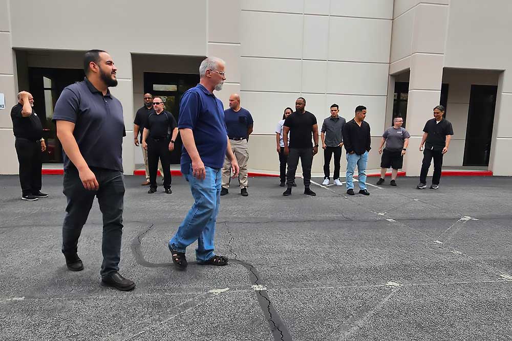 A group in casual dark clothing participates in executive protection training by Alexios Security Solutions near a beige building, as shown in "training-carousel-1"—two men walk ahead while others converse nearby.