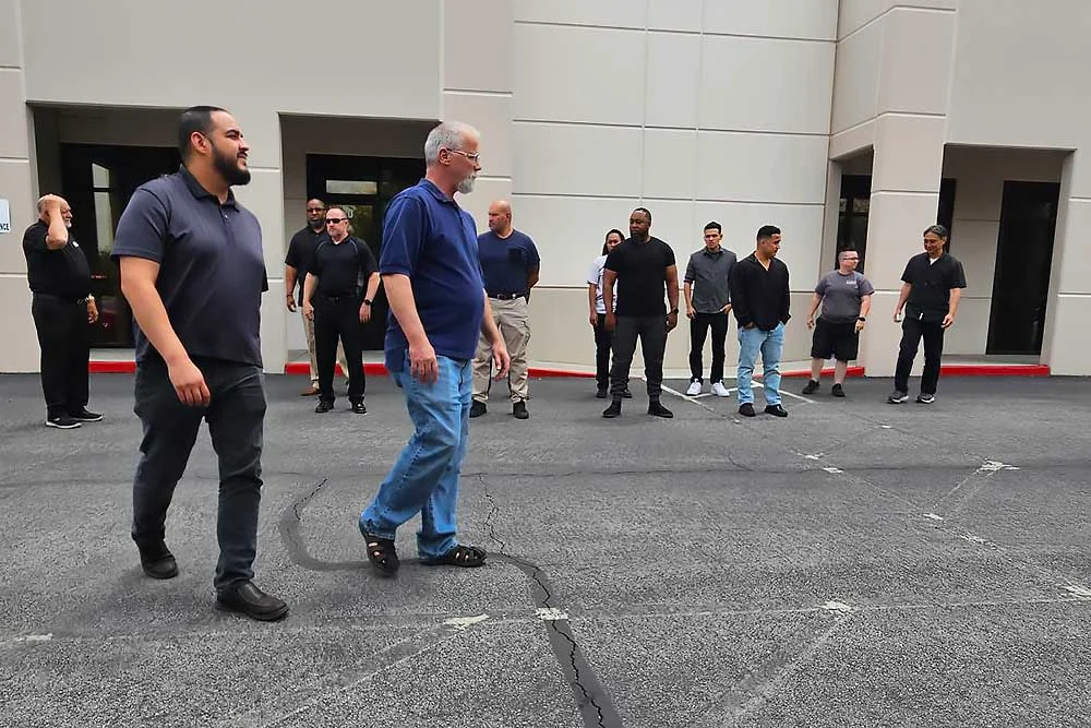 A group in casual dark clothing participates in executive protection training by Alexios Security Solutions near a beige building, as shown in "training-carousel-1"—two men walk ahead while others converse nearby.