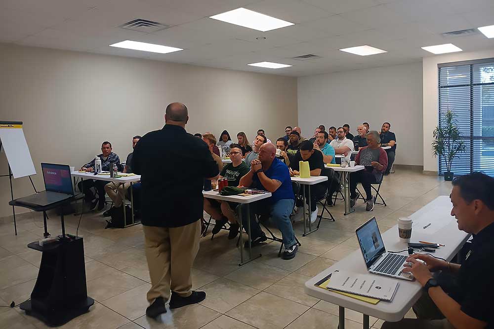 A man leads a behavioral intelligence training using the training-carousel-2 product, presenting to adults in a classroom with laptops, papers, and drinks. A flip chart and screen display materials for Alexios Security Solutions.