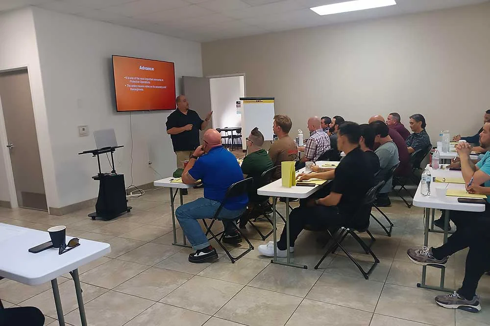 A man leads a training-carousel-3 executive protection session in Nevada, presenting to adults seated at tables with a nearby screen and whiteboard.