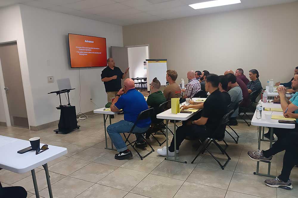 A man presents a behavioral intelligence training using training-carousel-5 to a group of adults in a classroom. A slide is projected behind him as attendees listen attentively, some taking notes.