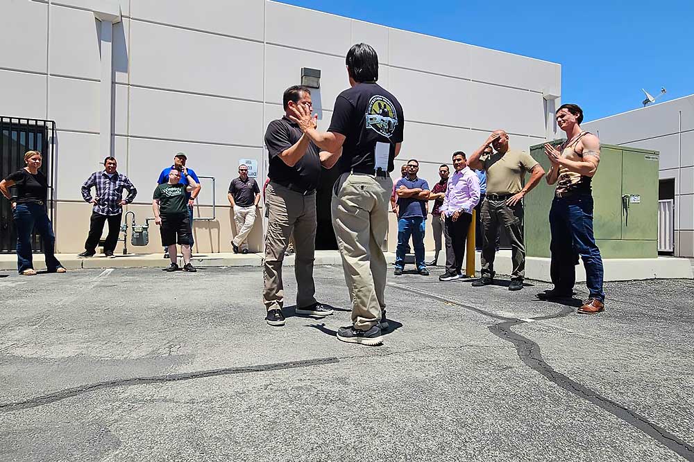 A group participates in Alexios Security Solutions' training-carousel-6, watching two men demonstrate a technique in a parking lot near a building under a clear blue sky.