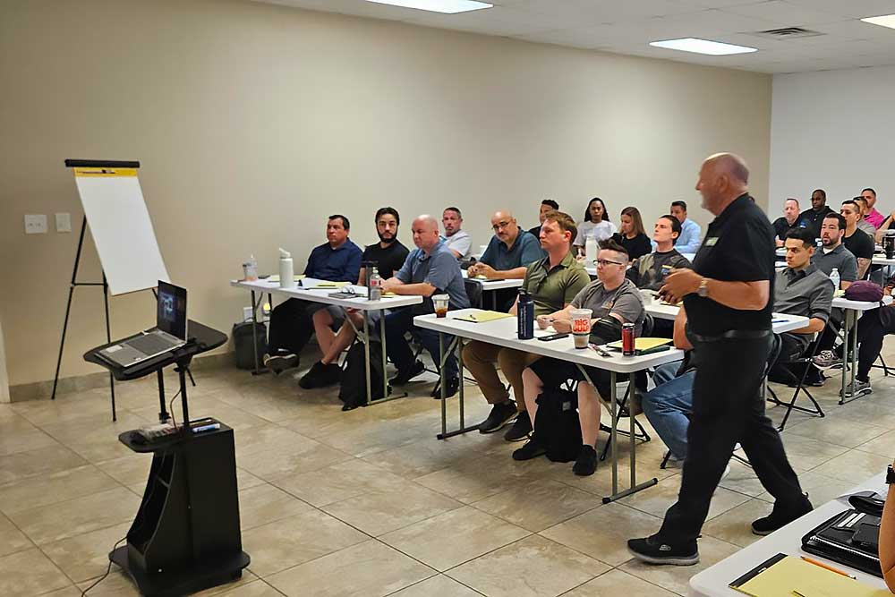 Adults attend a training session, seated at desks with notebooks and drinks, while an instructor presents at the front. The scene showcases the "training-carousel-7" by Alexios Security Solutions.