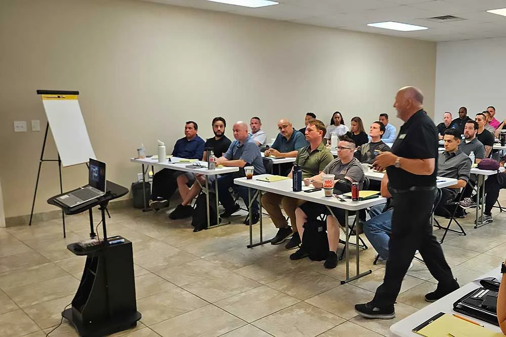 Adults attend a training session, seated at desks with notebooks and drinks, while an instructor presents at the front. The scene showcases the "training-carousel-7" by Alexios Security Solutions.