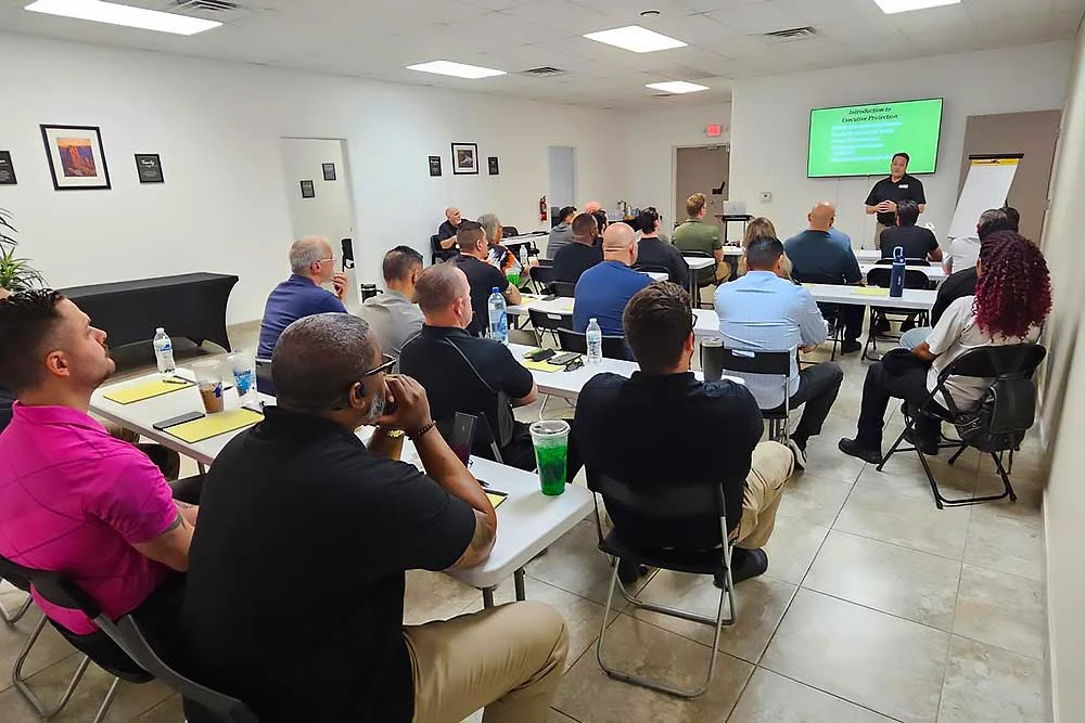 Adults attend a behavioral intelligence session (training-carousel-8), seated at desks with notebooks and drinks, listening to an instructor in front of a screen. Framed art decorates the classroom walls.