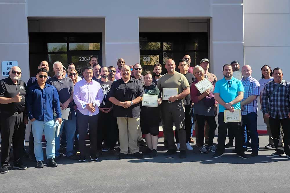 A group of men and women in business casual attire stand outside a building, celebrating the completion of training from Alexios Security Solutions and smiling at the camera while holding certificates.