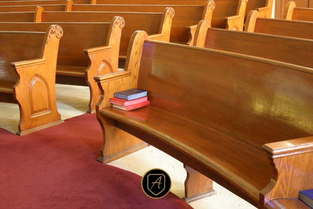 A few closed books rest on an empty wooden church pew, surrounded by rows of vacant seats and red and tan carpet. Product: The-Importance-of-Worship-Security-in-Nevada-Communities-2. | Executive Protection & Bodyguards in Las Vegas | Alexios
