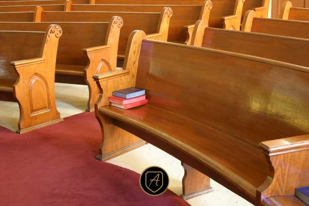 A few closed books rest on an empty wooden church pew, surrounded by rows of vacant seats and red and tan carpet. Product: The-Importance-of-Worship-Security-in-Nevada-Communities-2. | Executive Protection & Bodyguards in Las Vegas | Alexios