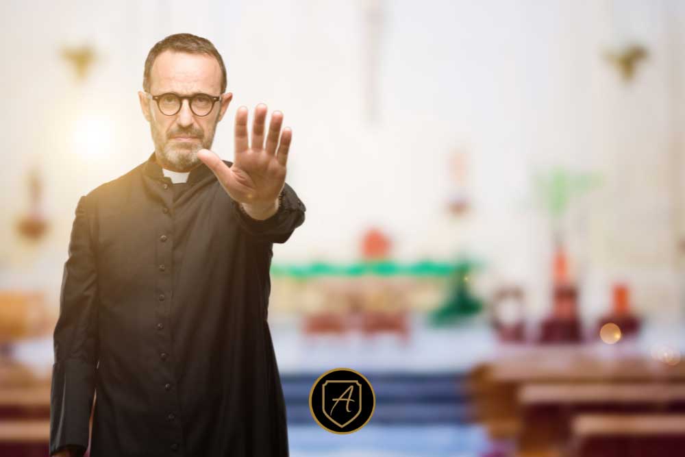 In "The-Importance-of-Worship-Security-in-Nevada-Communities-5," a priest in black robes stands in a church with his right hand raised, signaling stop. The blurred background reveals pews and an altar. | Executive Protection & Bodyguards in Las Vegas | Alexios